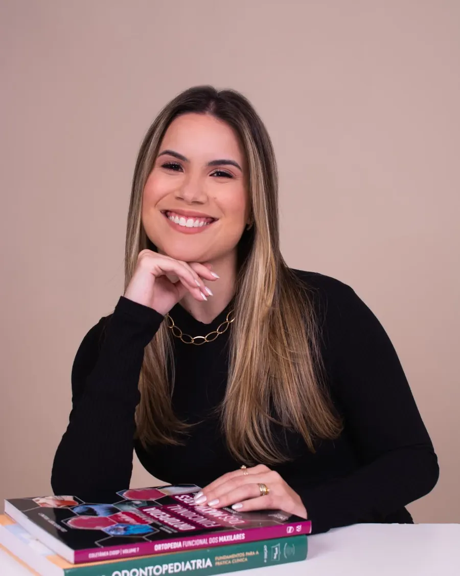Dra. Carolina Toledo sorrindo, apoiada em livros de odontopediatria